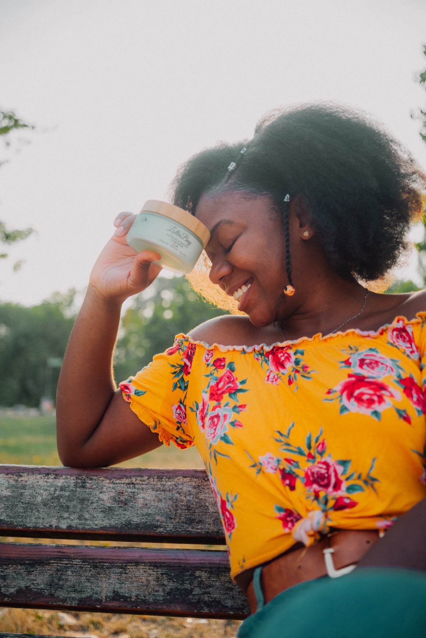A LolluBay customer holding the Nourish & Renew Hair Mask in golden-hour light on a park bench.